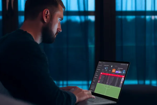 একজন পুরুষ তার ল্যাপটপে মনোযোগ দিয়ে কাজ করছেন, ব্যাকগ্রাউন্ডে লাইভ স্ট্যাটস — যা HHBD মোবাইল অ্যাপের স্পোর্টস ও গেমিং ফিচারগুলোকে তুলে ধরে।